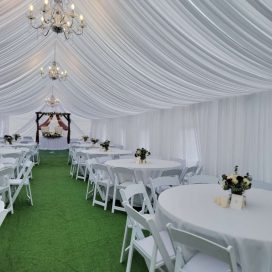 Elegant white draped wedding tent interior with chandeliers, round tables, and a green carpet aisle.