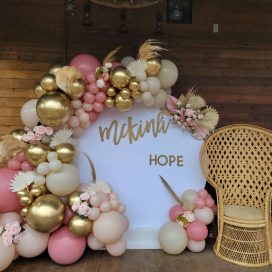 A balloon and flower arch with gold and pink balloons, surrounding a sign reading "McKinnah HOPE" next to a wicker chair.