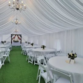 Elegant wedding tent interior with draped ceiling, chandeliers, white tables, chairs, and green floor.