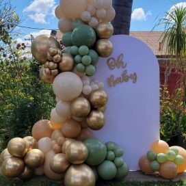 A photo of a festive "Oh Baby" sign adorned with clusters of green and gold balloons outdoors.