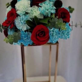 A bouquet of red roses and blue carnations atop a golden stand against a white backdrop.