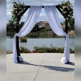 A wedding arch with white flowers overlooking a serene lake and residential backdrop.