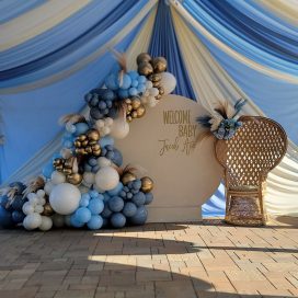 Baby shower decoration with blue and cream drapes, a balloon arch, and a wicker peacock chair.