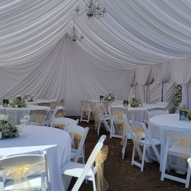 Elegant outdoor wedding tent with white drapery, chandeliers, and round tables with white chairs.