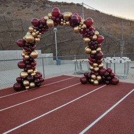 A balloon arch in maroon and gold colors stands on a running track with a hill in the background.