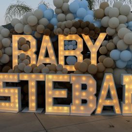 Illuminated "BABY ESTEBAN" sign adorned with a balloon arch at dusk.