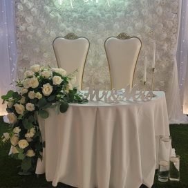 Elegant wedding table setup with "Mr & Mrs" neon sign, white floral backdrop, and draped lights.